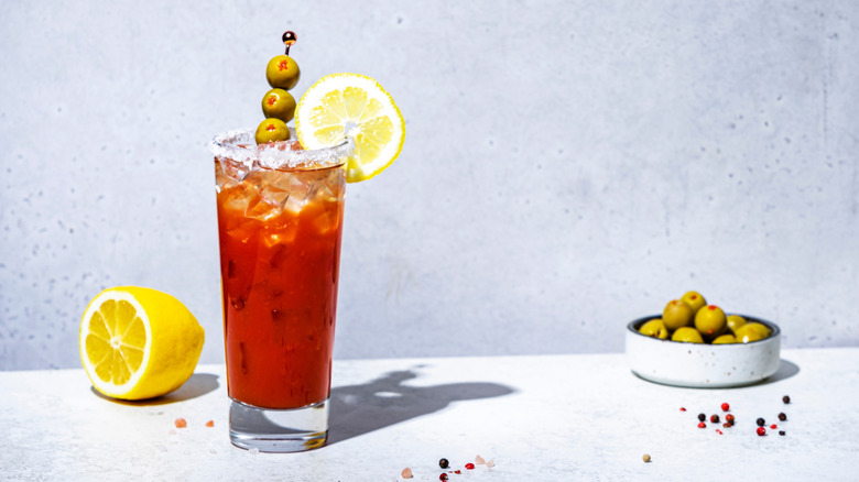 tomato juice-based drink in tall glass with olives and lemon garnish on marble table and grey background