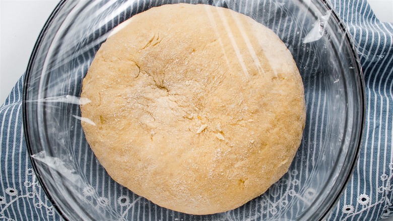 A bowl of pasta dough inside a glass bowl covered with plastic and placed on a blue kitchen towel