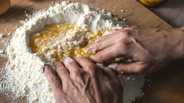 Two hands mixing eggs and flour to make pasta dough on a wooden pasta board