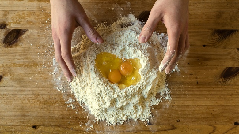 Two hands prepping pasta dough with flour and eggs on a wooden surface