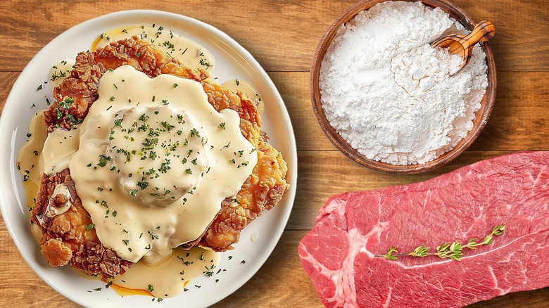 Plate of chicken fried steak with gravy next to flour in a bowl and a cut of steak on a wooden table