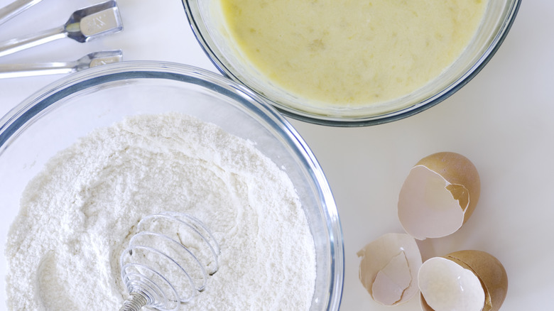 A bowl of flour and a bowl of wet batter with egg shells on the side