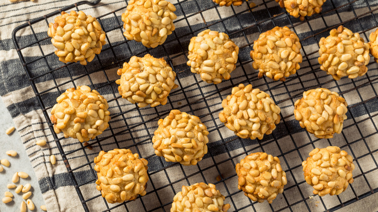 Italian pignoli cookies covered in pine nuts assembled on a cooling rack that is placed on a grey and white kitchen towel