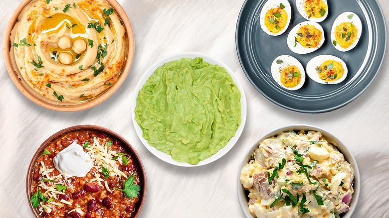 Composite image of various dishes with guacamole in the middle surrounded by hummus, chili, deviled eggs, and potato salad