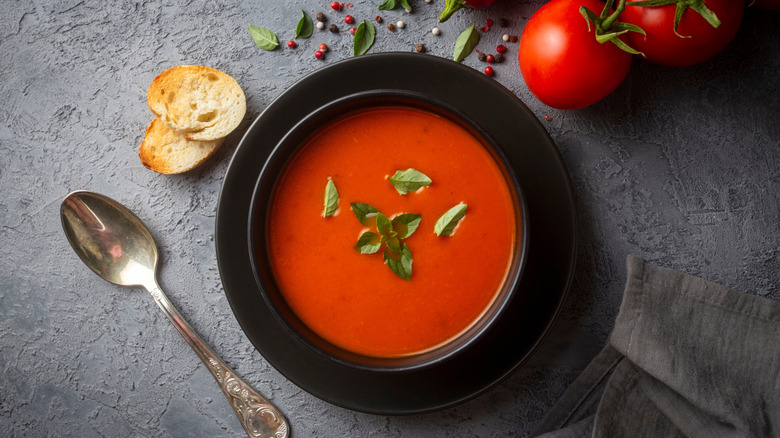 Tomato soup in black bowl next to fresh tomatoes and small toasts