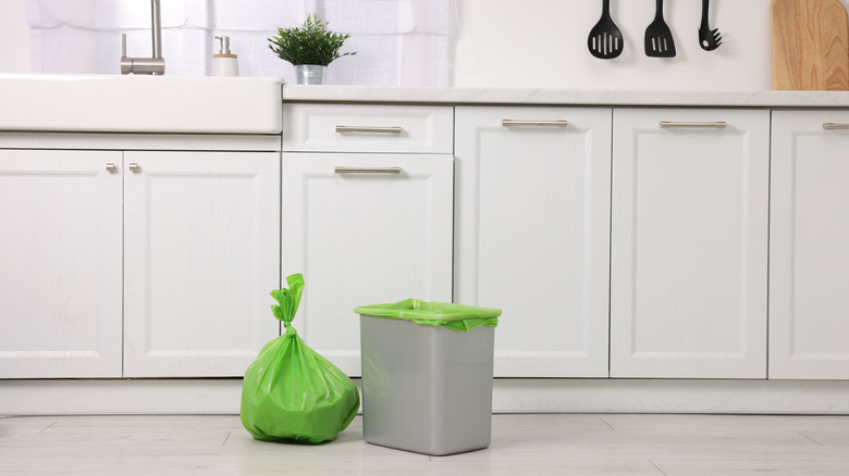 Plastic bag of garbage sitting beside trash can in a white kitchen