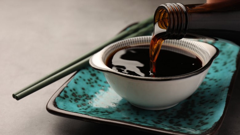 bottle pouring soy sauce into white bowl on blue plate with grey background