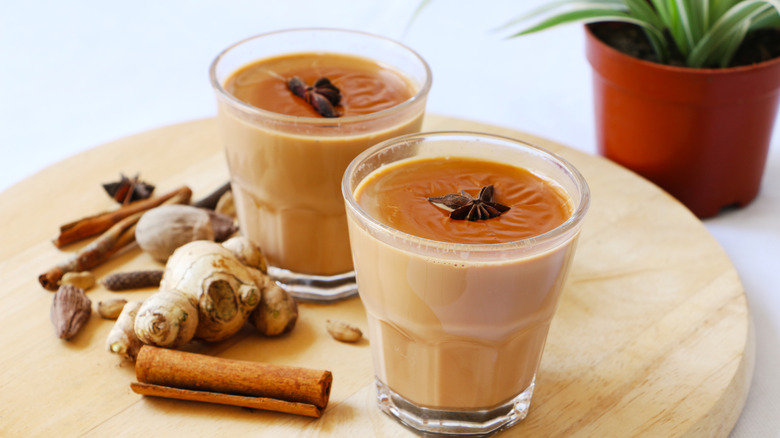 Two glasses of chai placed next to whole spices on a wooden platter and a plant in the background