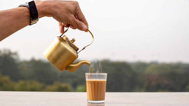 Hand pouring masala chai from a kettle into a glass