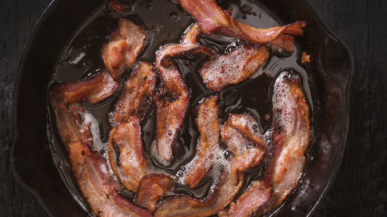 Bacon strips frying in a black cast iron pan drowned in sizzling fat