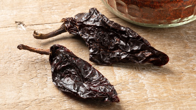 Two dried and smoked chipotle peppers on a wooden background with chipotle chili powder in the back