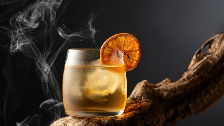 Cocktail filled with ice and garnished with an orange wheel sitting on wooden branch surrounded by smoke and a dark background