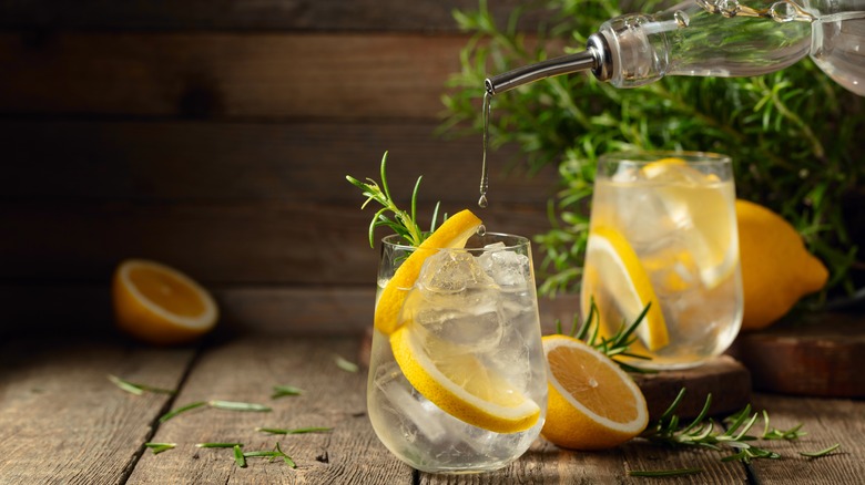 Pouring clear liquid into a cocktail glass filled with ice, lemon slices, and rosemary that is placed on a wooden table, with another glass, rosemary, and lemon halves in the background