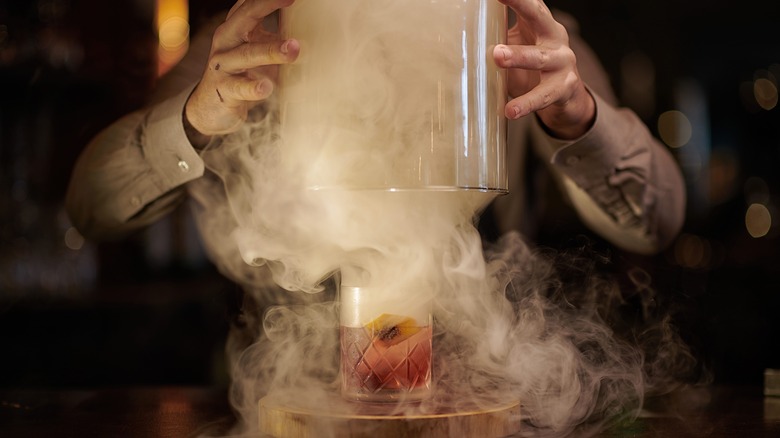 Bartender covered in smoke, lifting a glass cover off a Negroni cocktail that's been exposed to smoke