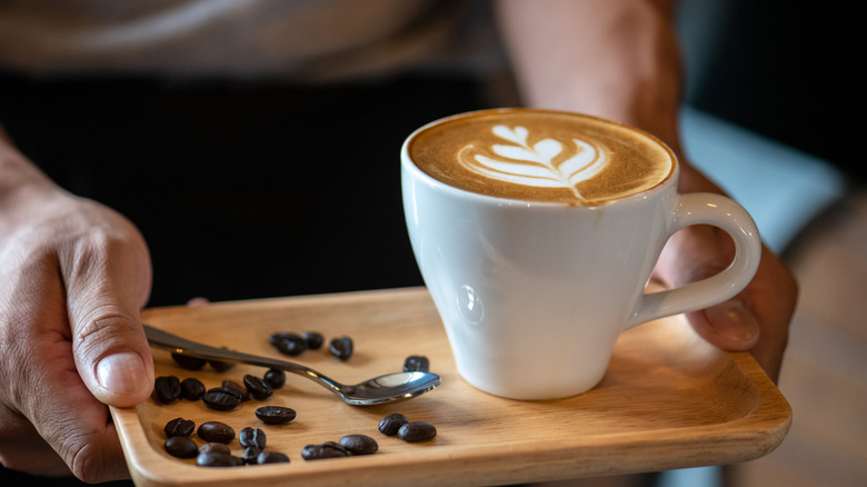 A closeup shows a barista bringing out a tray with coffee that has art on top.