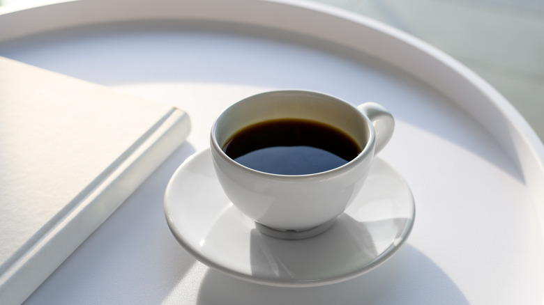 A black coffee fills a white cup on top of a saucer that's on a white table.