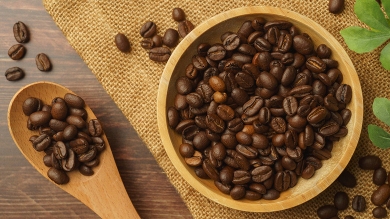 Coffee beans fill a wooden bowl that is next to a wooden spoon.