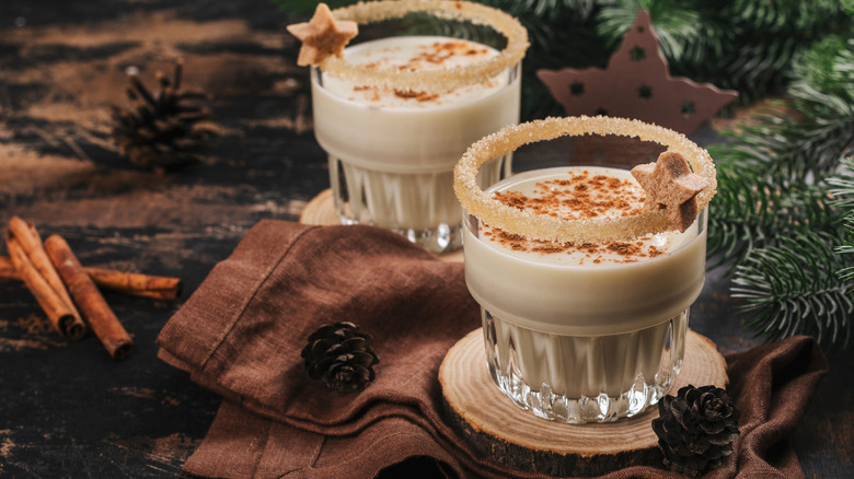 Two glasses of eggnog with festive garnishes are arranged in a rustic setting.