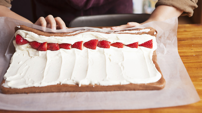 Hands rolling up sponge cake filled with cream and strawberries