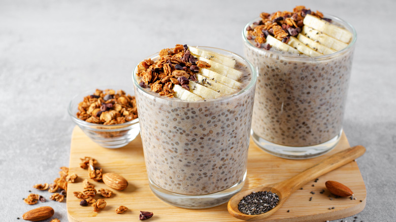 Two cups of chia seed pudding with banana and granola on wooden cutting board