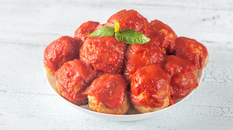 Bowl of saucy meatballs in white bowl with basil leaf on top