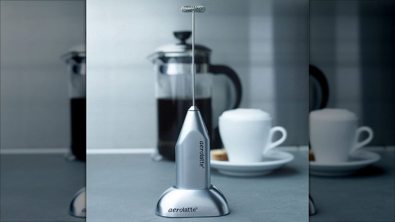 Silver Aerolatte milk frother on stand with coffee cup in background