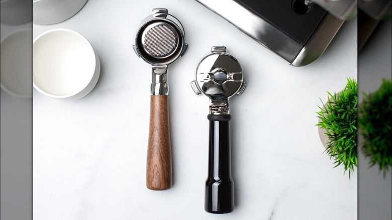 Two Crema portafilters next to one another on countertop, one wooden and one black