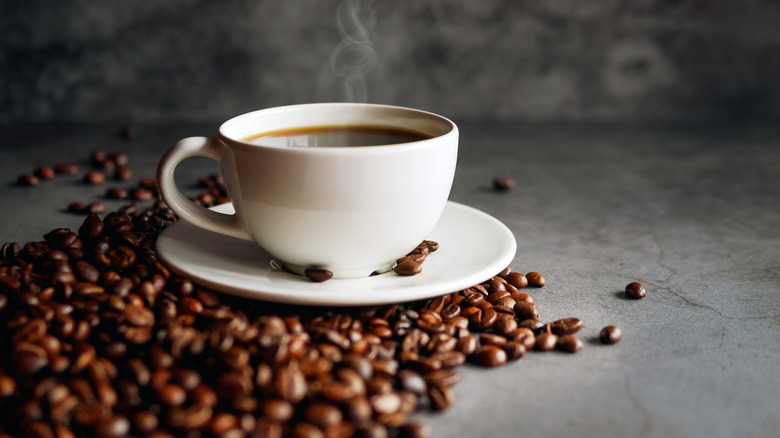 White cup of coffee on saucer and coffee beans on grey surface