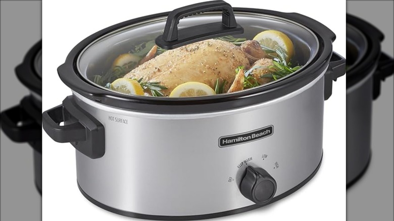 large, silver slow cooker on white background with whole chicken, lemon, and rosemary inside