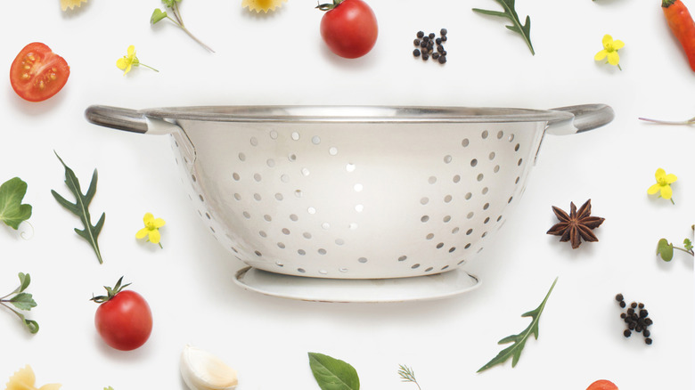 White colander on white background surrounded by fruits, greens, and herbs
