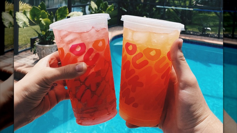 Two hands holding Dunkin' Refreshers in front of a pool