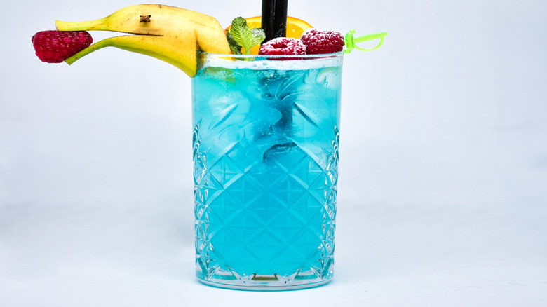 A blue tiki cocktail with garnished with raspberries, mint, orange slice, and a banana dolphin that is holding a raspberry in its rostrum