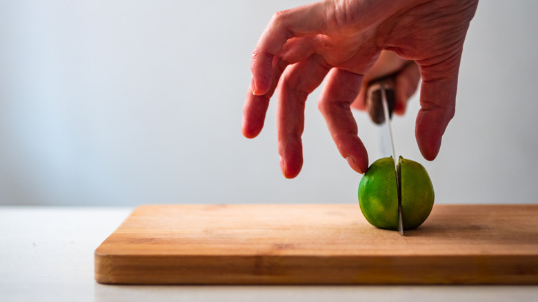 A person using a knife to slice a lime in half on a wooden cutting board