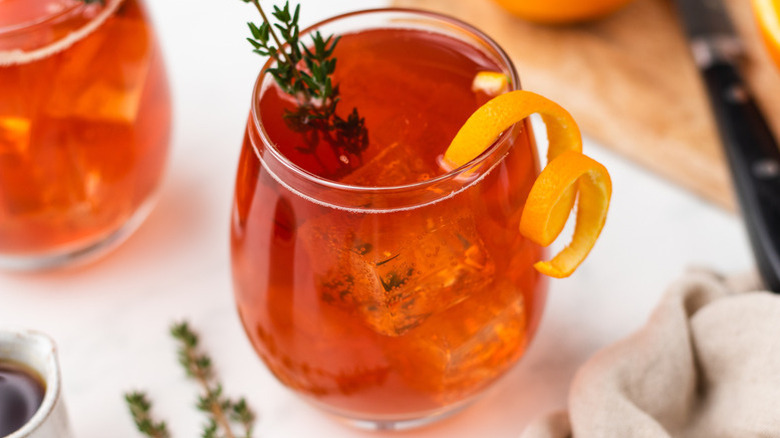 A close-up of an orange cocktail packed with ice and garnished with thyme sprigs and an orange twist