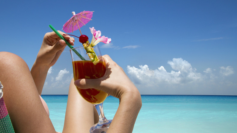 A person on the beach holding a cocktail garnished with a cocktail umbrella, flower, maraschino cherry, and a pineapple wedge