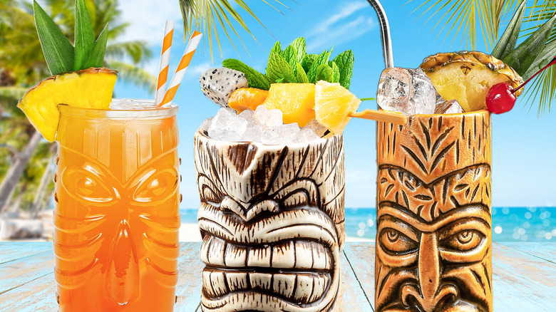 Three tiki-style cocktails with various garnishes lined up on a table with a beach scenery in the back