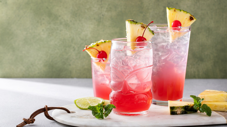 Three pink tiki cocktails garnished with Maraschino cherries and pineapple wedges lined on a tray and surrounded by lime, mint, and pineapple