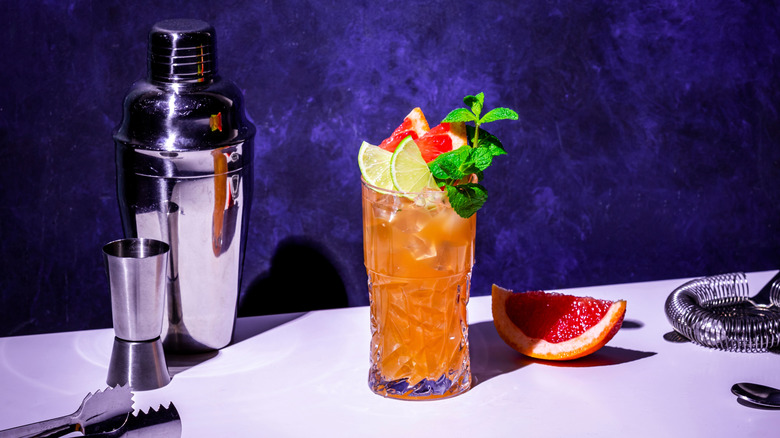 Zombie cocktail garnished with mint leaves, lime, and blood orange resting on a white counter and in front of a blue wall surrounded by classic cocktail shaker and a citrus wedge