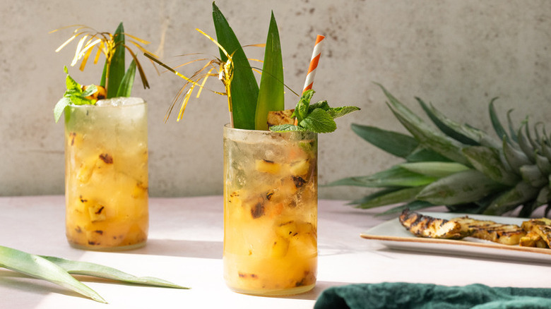 Two pineapple cocktails in tall glasses garnished with pineapple fronds set on a beige background next to a pineapple crown