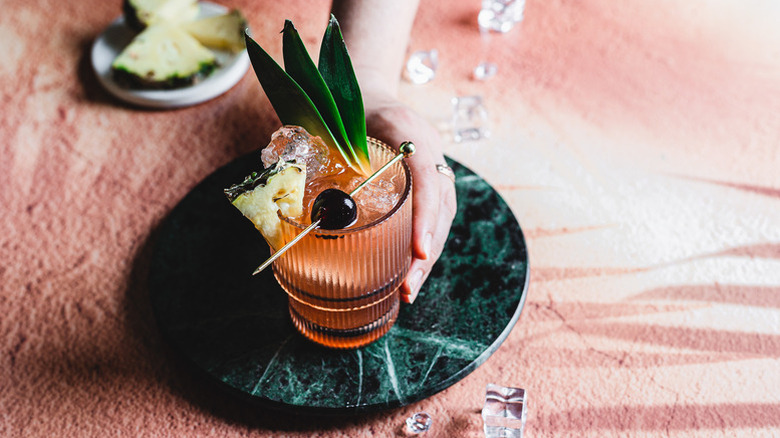 A hand holding jungle bird cocktail packed with ice and garnished with a Luxardo cherry, fresh pineapple, and pineapple fronds