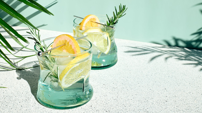 Two summery cocktails with rosemary sprigs and pierced lemon slices lined on a white counter surrounded by green leaves