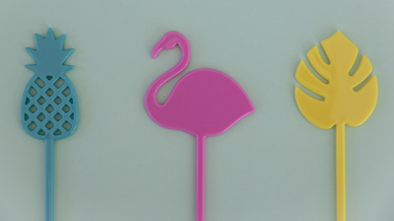 Three colorful tropical-themed cocktail swizzle sticks made of plastic depicting a pineapple, flamingo, and a leaf