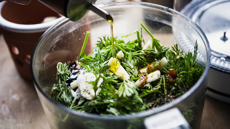 Food processor full of herbs with a stream of oil pouring in