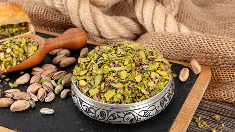 Silver bowl of chopped pistachios with whole pistachios and wooden scoop