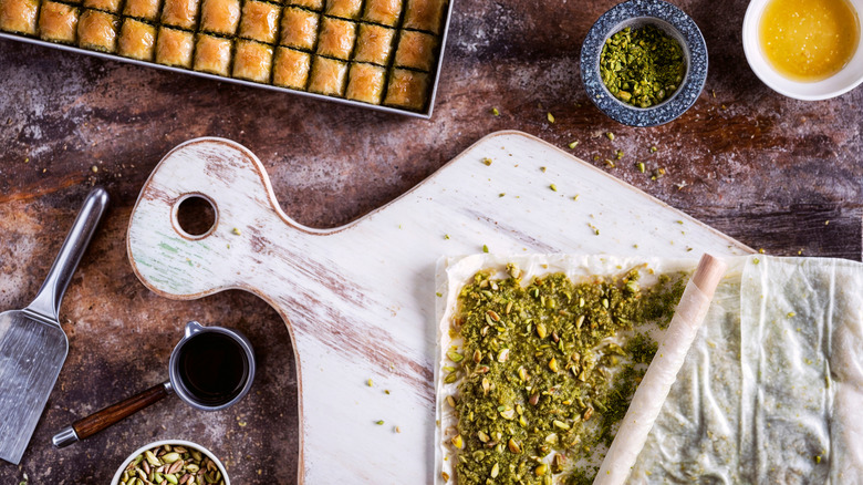 Making baklava on wooden chopping board with baked baklava in tray and pistachios in bowls