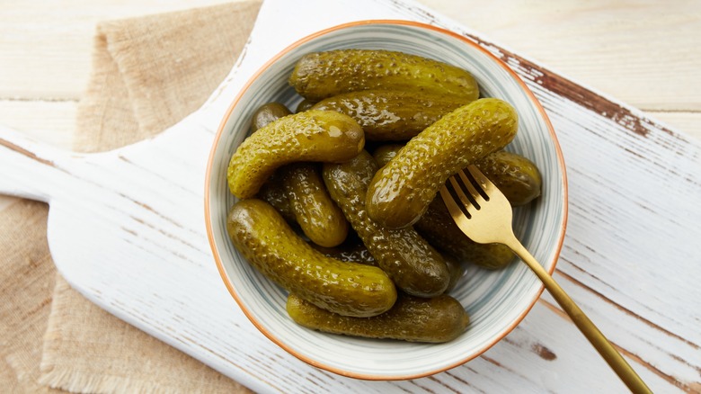 Small cornichon pickles with a fork in a ceramic bowl on a white wooden board