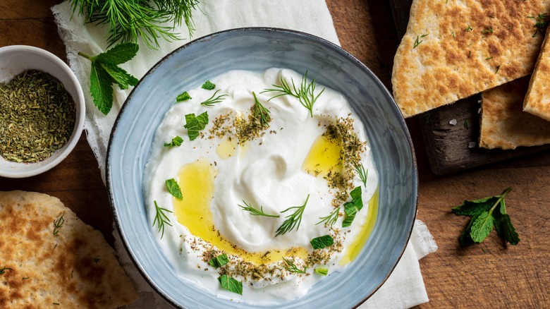 Labneh spread in a blue plate garnished with herbs, olive oil and spices surrounded by pita bread, fresh herbs, and dry spices