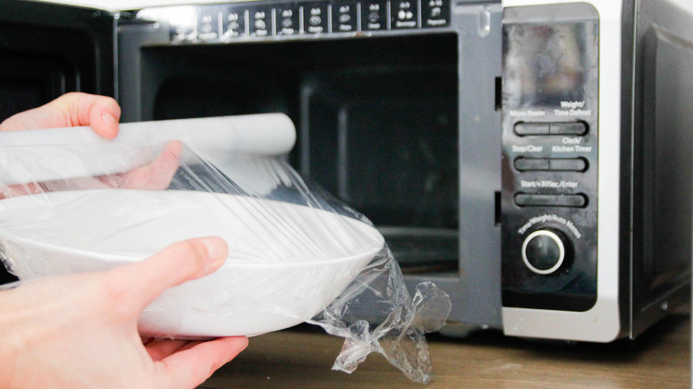 covering dish in plastic wrap in front of microwave