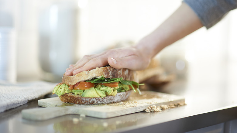 One hand pressing down sandwich with vegetables and avocado