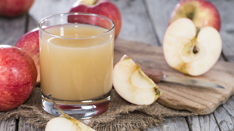 Glass of cloudy apple juice on a jute rag with whole, sliced, and halved apples in the back